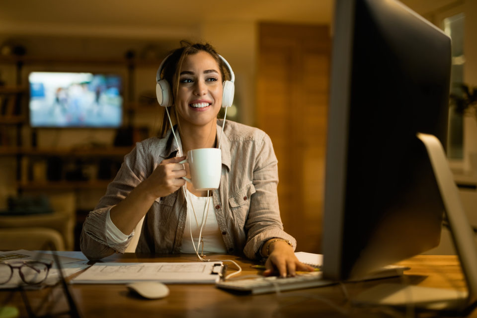 Happy woman with headphones surfing the net on desktop PC at night.