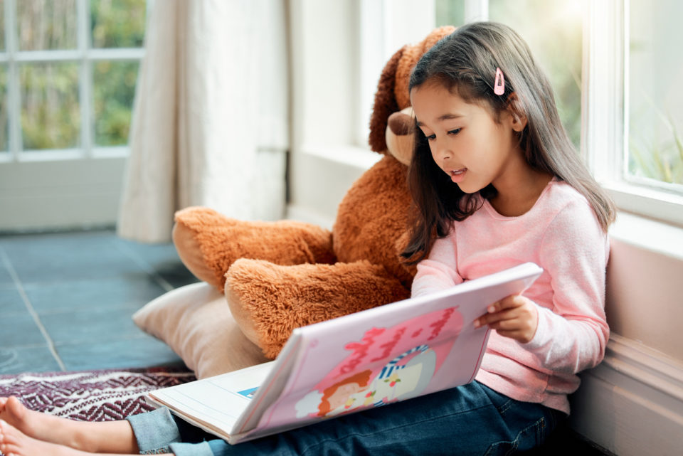 A little girl in a pink shirt, reads a book under a floor-to-ceiling window